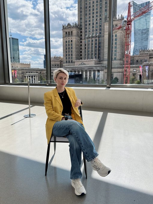 A woman with short blonde hair wearing a yellow blazer and jeans sitting on a chair in front of large windows overlooking a city with historic buildings.