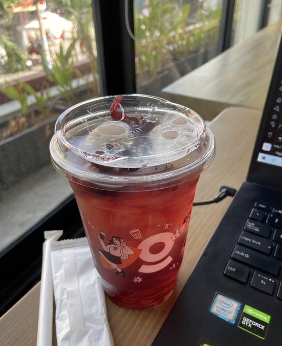 A plastic cup of iced red drink sits next to a laptop and white napkins on a café table.