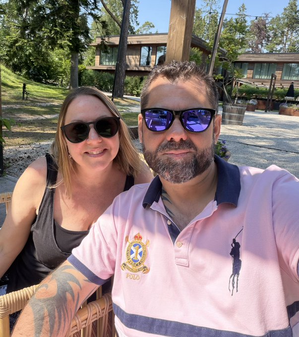 The image features a man wearing sunglasses, a pink polo shirt with a gold emblem, and a woman sitting next to him. They are both smiling for the camera. The caption of the photo reads "23 years married today! Look how happy she is..its because she doesnt know how much money I lost in NFTs". This post seems to be about an event or celebration involving the couple, as they appear to be enjoying their time together and sharing a moment captured in the image.