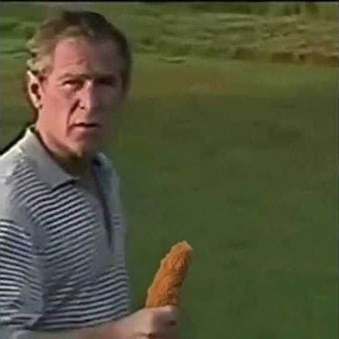 A still of a man with a serious expression wearing a striped polo shirt, holding what appears to be a fried snack, with a grassy background.