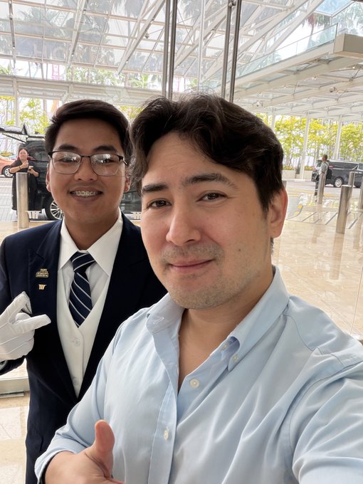 A man in a light blue shirt takes a selfie with a smiling hotel valet wearing white gloves and a dark suit with a striped tie, inside what appears to be a luxury hotel entrance with glass walls and vehicles visible outside. The post highlights the man's chance encounter with "Ian," an Axie Infinity player whose story reflects the post's theme of meaningful connections made during the pandemic era of crypto gaming.