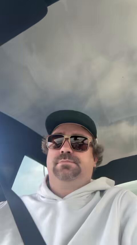 The image features a man wearing sunglasses, a white hoodie, and a baseball cap while sitting inside a car. The man appears to be looking out of the window or at someone outside the vehicle. There are no texts or other objects visible in the photo. The caption provided seems unrelated to the main subject matter and does not provide any additional information about the scene depicted in the image.