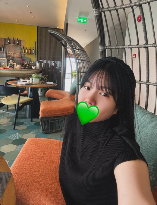 A selfie of a woman in a black top with a green leaf emoji over her mouth, taken in a colorful restaurant/lounge setting.