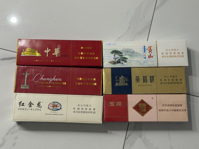 Six packs of Chinese cigarettes laid on a tiled floor, with brands such as 'Chunghwa FILTER CIGARETTES', '黄鹤楼', '红金龙 HONGJINLONG', '玉溪', '黄山', and '中华' visible, along with health warning labels.