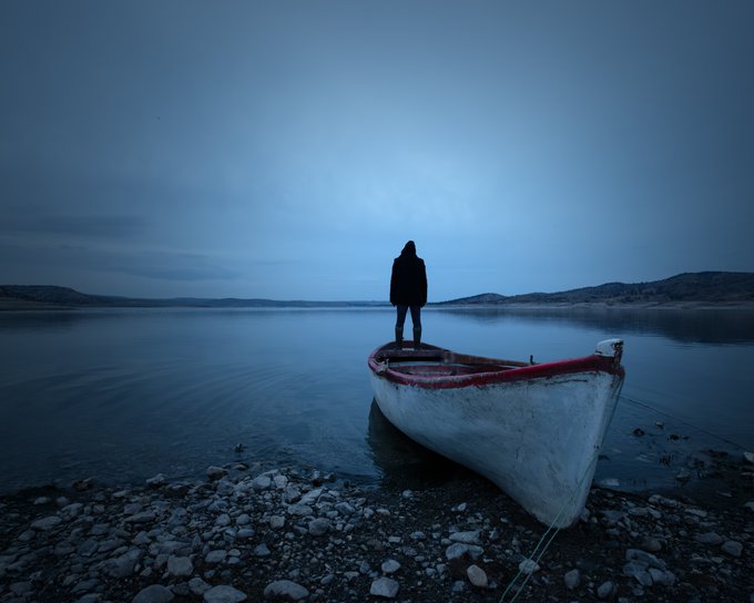 A silhouetted figure stands on a small white and red boat moored at a rocky shore, gazing out over a calm lake under a moody blue twilight sky.
