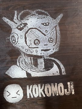 A rustic, monochromatic illustration features a stylized cartoon character resembling an astronaut or robot with horns, drawn in what appears to be salt or grain art on a dark surface, accompanied by the text "KOKOMOJI" and a small emoticon face "><". This corresponds to the accompanying post, which mentions it is salt art created for the @Kokomojinft account, blending pixelated digital art aesthetics with the textured, granular quality of salt.