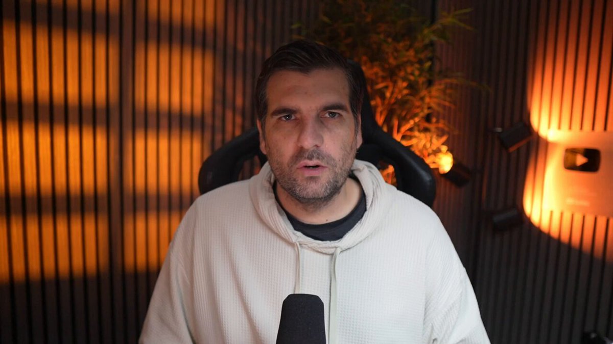 The image shows a man wearing a white hoodie sitting behind a microphone with his hands on his knees. The background features a dark wall, which contrasts with the bright light shining through from above. There are also two lights visible in the scene - one located near the top left corner and another towards the bottom right corner of the frame. The image is related to a post discussing Wall Street's massive bet on digital assets, specifically XRP (Bitcoin Cash), and how it has changed the narrative surrounding hidden institutional capital flows.