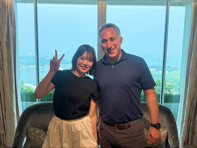 Two people pose for a photo in front of a window; a younger woman in a black top and beige skirt makes a peace sign while standing beside a man in a navy polo shirt.