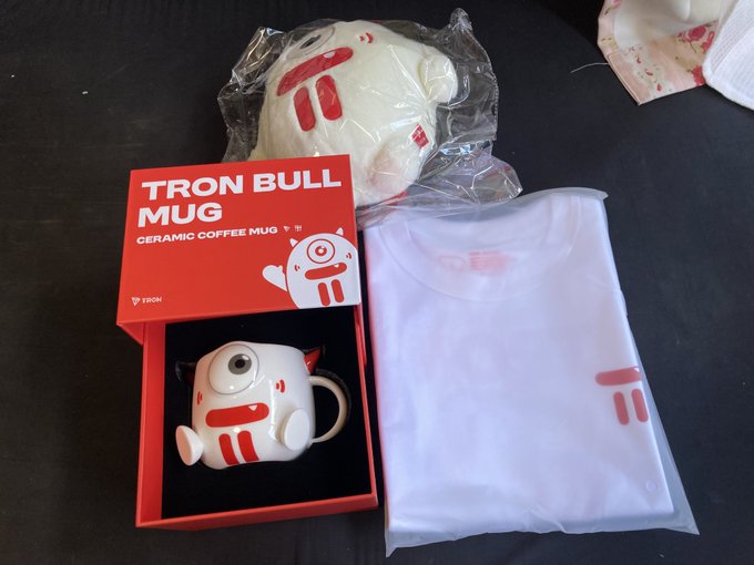 A photo of a 'TRON BULL MUG' ceramic coffee mug in a red box, shown alongside its matching white T-shirt in a plastic bag on a dark surface.