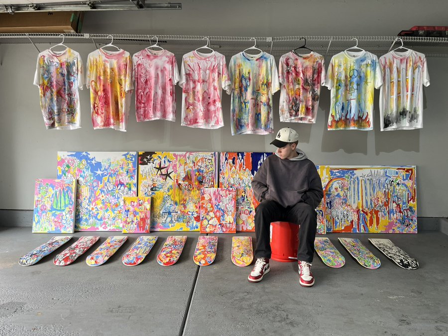 A man wearing a cap, brown sweatshirt, and red sneakers sits on a red stool in a garage displaying colorful, abstract painted t-shirts, skateboards, and canvas paintings.