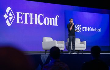 The image shows a man standing at a podium with a microphone, giving a speech or presentation. The room has blue walls and two white chairs placed near the stage. There are several people sitting in the audience, attentively listening to the speaker. A large screen is visible behind the speaker, displaying the name "ETHConf" on it. The image also includes text that reads: "What a time. Thank you for being part of ETHConf. Three days of big ideas, packed rooms, and conversations that will shape what'S next for Ethereum."