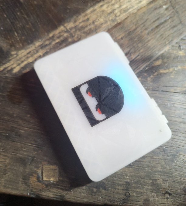 A small 3D-printed Batman-themed Darth Vader helmet sitting on a white base with a blue glow, displayed on a wooden surface.