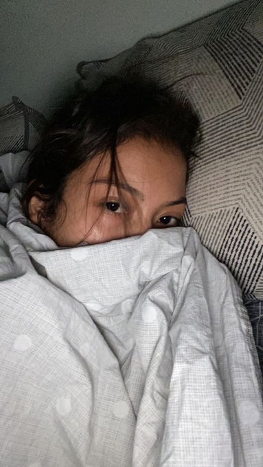 A young woman lying in bed, peering over a light grey patterned duvet, with only her eyes and forehead visible.