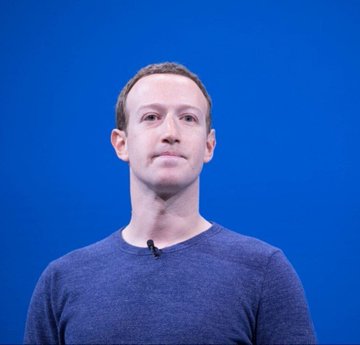 A man with a serious expression stands against a bright blue background, wearing a dark blue sweater with a small lapel microphone visible. The image likely depicts Mark Zuckerberg, CEO of Meta, in connection with the news about Meta launching its new consumer AI agent platform "Hatch."