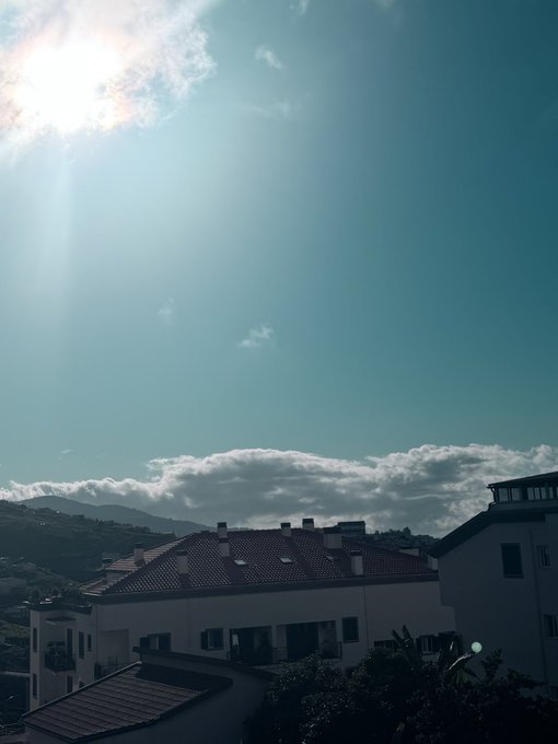 A sun-drenched view from a rooftop captures a red-tiled residential building with solar panels, set against a backdrop of distant mountains and a bright blue sky with scattered clouds. The scene, tagged with "GM" and a request for a SOL (Solana) address, evokes a sunny morning greeting—a common crypto community ritual—while the solar panels subtly tie to the Solana network's name.