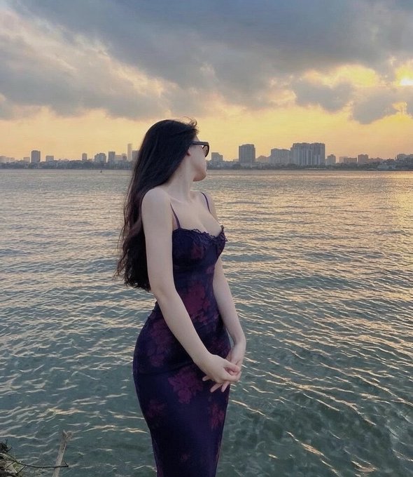 A woman in a floral purple dress and sunglasses stands by the waterfront at sunset, gazing toward a city skyline in the distance.