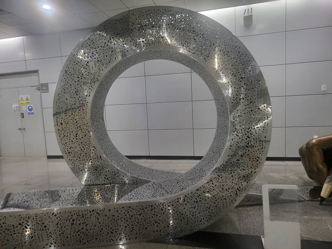 A large, modern circular metal sculpture with intricate cut-out patterns stands in what appears to be a building lobby or public space.