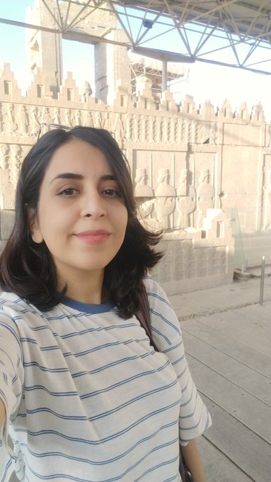 A woman in a white and blue striped shirt takes a selfie in front of ancient stone carvings depicting figures in relief.