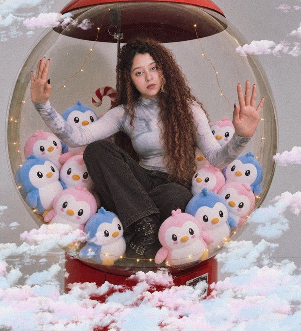 A young woman with long curly hair sits inside a clear spherical snow globe-like enclosure filled with pink and blue penguin plushies and fairy lights, surrounded by cotton-candy clouds.