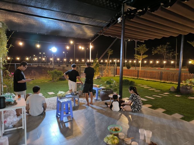 A nighttime outdoor gathering under a covered patio with string lights, where a group of people including adults and children are gathered around a table, with some cooking food and bowls of ingredients on the floor.