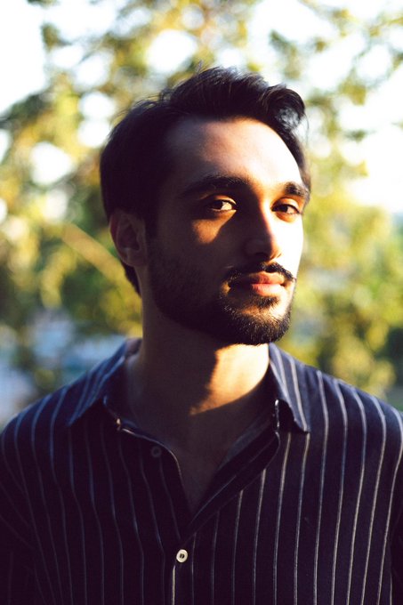 A portrait of a bearded man with dark hair wearing a dark striped shirt, standing outdoors with sunlight illuminating the side of his face.