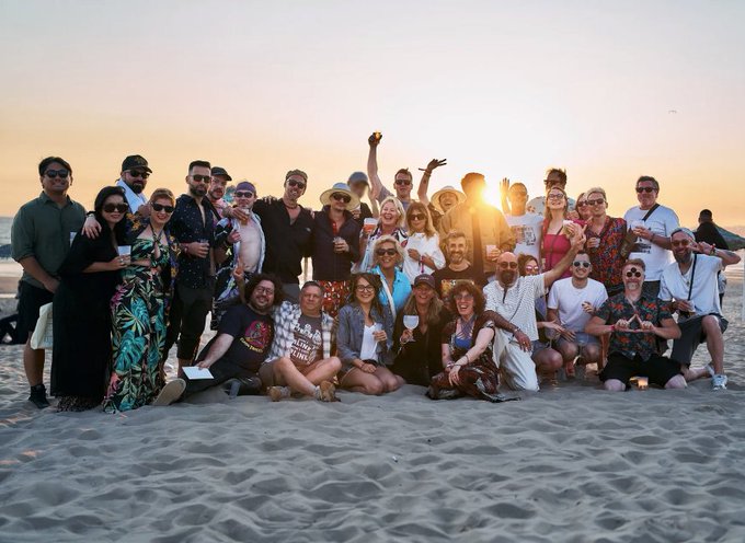 The image shows a group of people posing together for a photo at the beach during sunset. The sun is setting behind them, casting a warm glow over their faces as they gather around each other. There are at least 15 people visible in the picture, with some standing and others sitting on the sand. They appear to be enjoying themselves and capturing this moment of camaraderie.

The photo also contains text that reads "@NFCsummit is so much more than a conference. It'S a reunion of dreamers, artists, builders, collectors, and friends who somehow found each other through this crazy web3 phenomenon and then turned". This message adds an interesting layer to the scene, as it suggests that these people have come together for a special event or gathering related to cryptocurrency or blockchain technology.