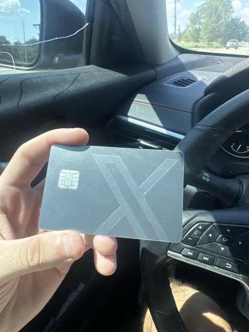 A hand holds a sleek, dark grey metallic card with a large 'X' design and an embedded chip, photographed inside a vehicle.