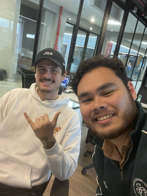 Two young men smile for a selfie in an office setting; the man on the left wears a white hoodie with a small bee logo and a black 'Jito' cap, making a hand gesture, while the man on the right wears a dark zip-up jacket.