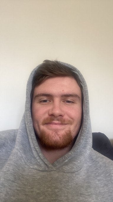 A young man with a ginger beard wearing a grey hoodie smiles at the camera against a plain wall, posted with "GM" for a casual good morning greeting.