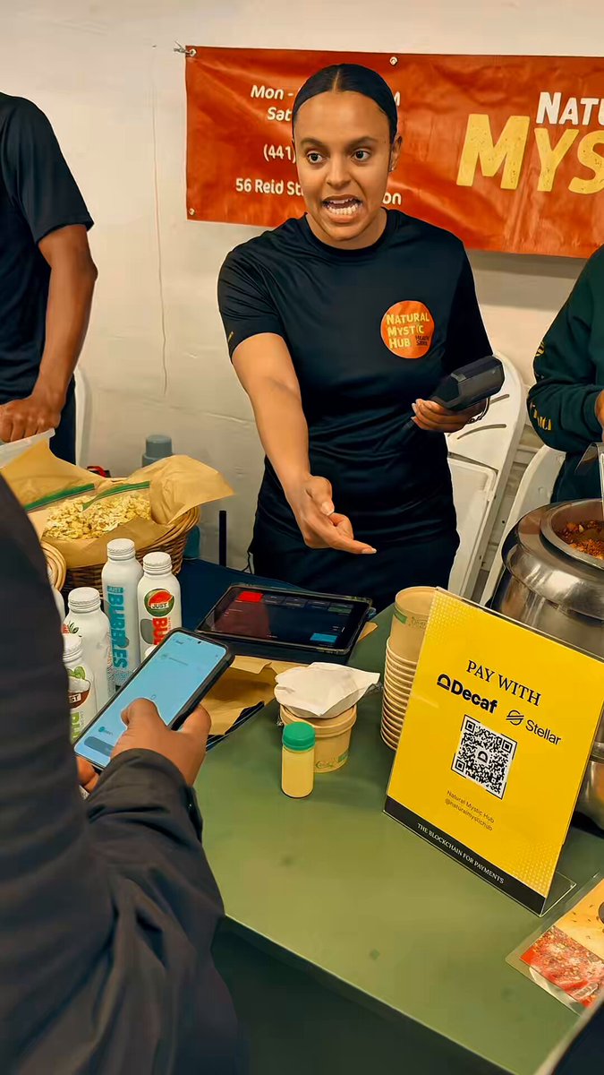 The image shows a woman standing behind a table, holding a cell phone up for a picture while another person looks at her. The background features an orange banner that reads "National MyS" along with some text about the restaurant's offerings. There are also two other people in the scene, one of whom is wearing a black shirt and appears to be looking at the woman taking the photo.