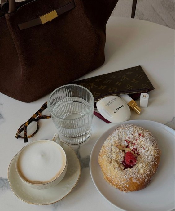 A flat-lay photograph of luxury items on a marble table, including a brown Hermès bag, Louis Vuitton pouch, Chanel hand cream, glasses, a cappuccino, and a raspberry pastry.