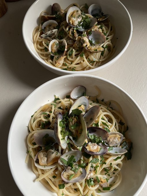 Two white bowls filled with spaghetti pasta topped with open clams in their shells, fresh chopped parsley, and garlic, served on a white table.
