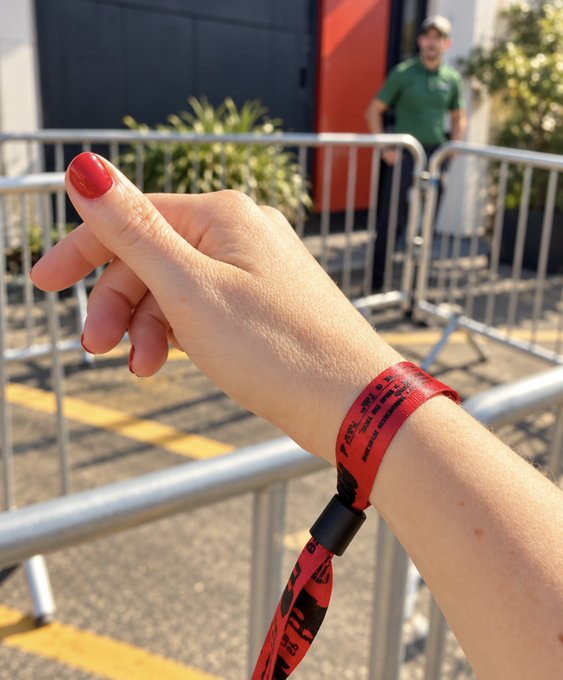 A first-person view of a hand with red nail polish wearing a red event wristband, with a person in a green shirt visible behind metal barricades.