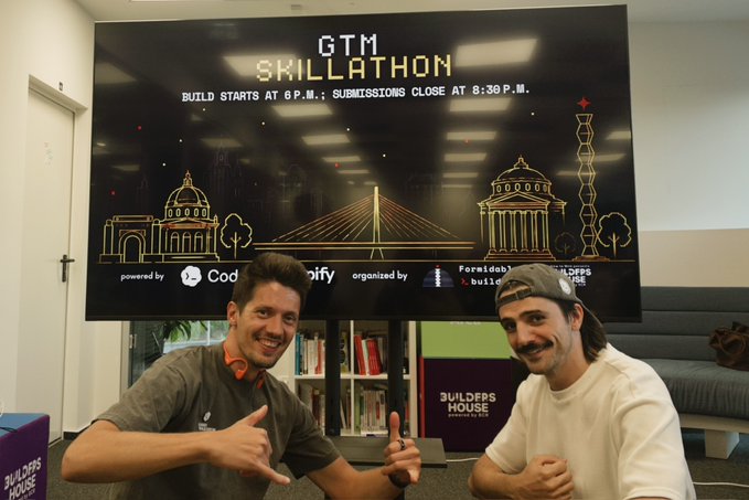 Two men sit in front of a black banner for a 'GTM SKILLATHON' event, with text indicating 'BUILD STARTS AT 8 P.M.; SUBMISSIONS CLOSE AT 8:30 P.M.'
