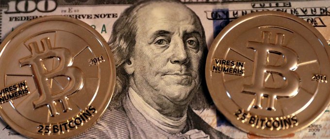 Two large, physical Bitcoin coins marked '25 BITCOINS' and 'VIRES IN NUMERIS' are displayed on top of a United States one hundred dollar bill featuring Benjamin Franklin.