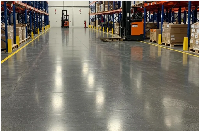 A polished concrete warehouse aisle flanked by tall pallet racks stocked with cardboard boxes, with yellow safety bollards lining the storage bays and a forklift visible in the distance near an open loading door. The image grounds the post's message by showing the real, operational "packing hall" — the tangible warehouse environment where the ~416 open bounties and ~$317k USDC in settled work are actually being executed, not a conceptual slide.