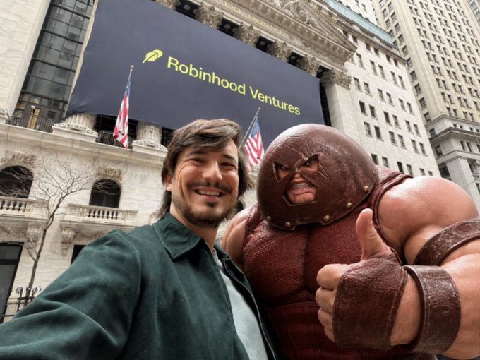 A man taking a selfie with someone in a Juggernaut (X-Men) costume outside the New York Stock Exchange, which has a 'Robinhood Ventures' banner displayed.