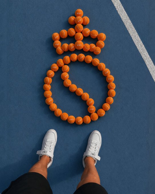 An overhead view showing a person's white sneakers standing on a blue tennis court next to tennis balls arranged in the shape of a money bag/dollar sign.