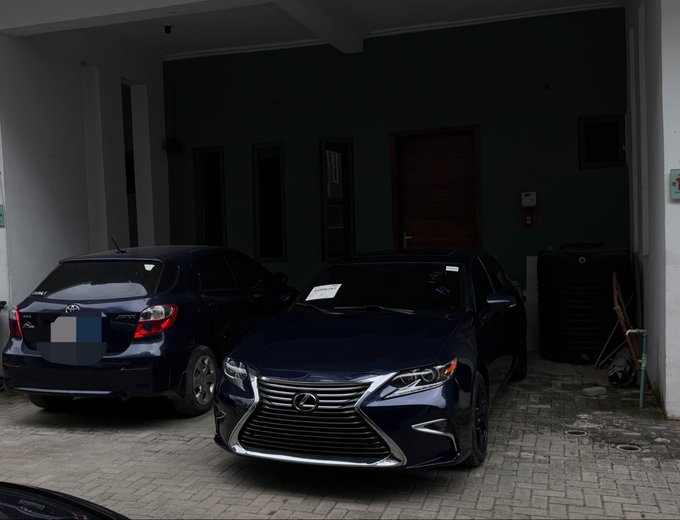 Two dark-colored sedans are parked in a covered driveway, with the prominently featured car in front being a Lexus (bearing the Lexus emblem and "LEXUS" badge on the grille). The vehicles sit in front of a building entrance with closed doors, and a piece of paper visible on the windshield appears to display text—though it cannot be clearly read—possibly related to the "gm." caption reference.