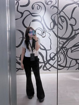 A mirror selfie of a woman wearing sunglasses, a white t-shirt, and black flared pants, standing in front of a wall decorated with abstract black line art.