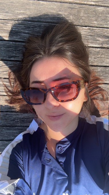 A selfie of a young woman with brown hair wearing tortoiseshell sunglasses and a blue collared shirt, taken against a wooden background in bright sunlight.