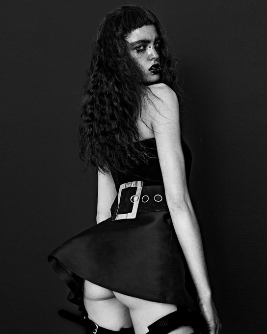 A black and white fashion photo features a woman with long dark hair posing in a black strapless dress with a large belt buckle against a dark background.