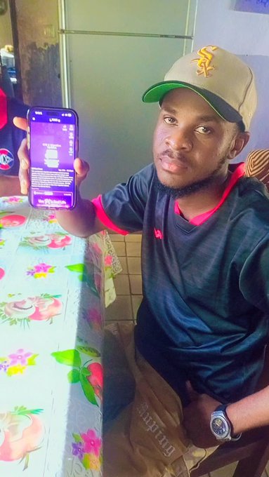 A young man wearing a beige cap with 'SF' lettering and a dark shirt with red trim takes a selfie while holding up a smartphone displaying an app interface, seated at a table with a floral tablecloth.