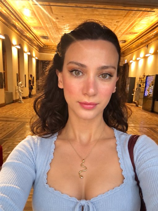 A woman with long dark hair poses for a selfie in an ornate, gilded hallway, wearing a light blue ribbed top and a gold snake pendant necklace. The elegant, glamorous photo complements her upbeat "Monday motivation" message to her Web3 community.