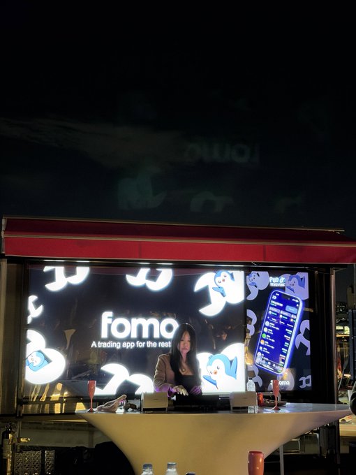 A woman poses behind a DJ booth at night with a large illuminated screen displaying the "fomo" trading app logo and the tagline "A trading app for the rest of us," featuring cartoon illustrations of dollar signs and a phone interface—visually capturing the "fomo world order" theme of crypto-trading culture.