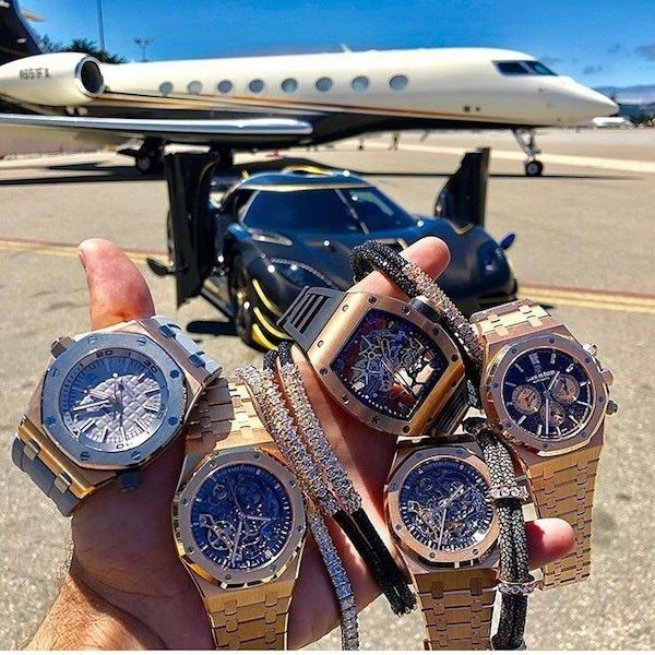 A close-up of a hand holding several luxury gold and diamond watches, with a private jet and a sports car parked on the tarmac in the background.