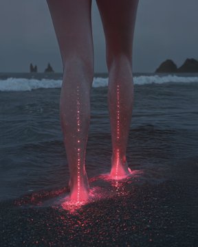 A person's legs stand at the edge of a beach at twilight, illuminated from below by bright red lights shining through the sand and water.
