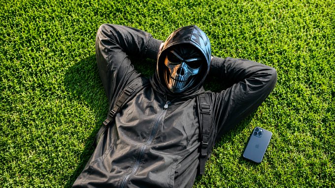 A person wearing a skull mask and black hooded jacket lies on bright green grass with their hands behind their head, a smartphone resting beside them.