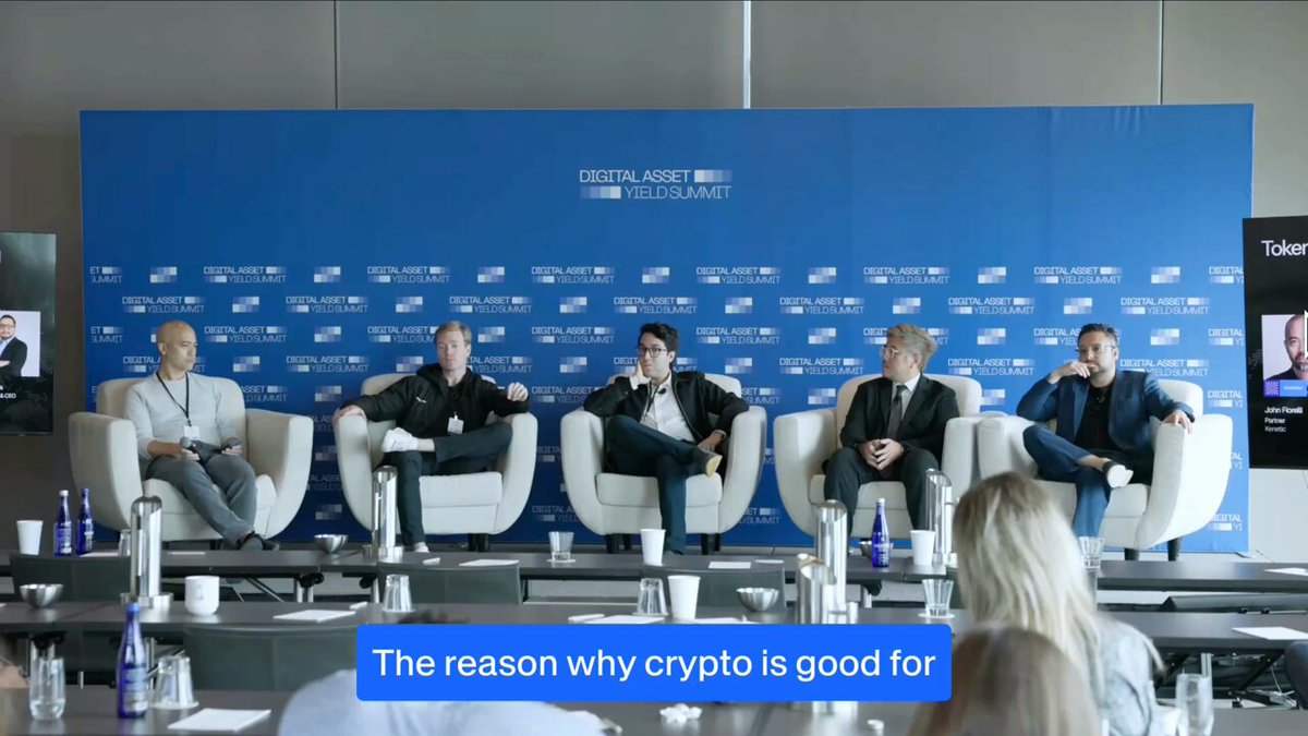 The image shows five people sitting around a table with chairs arranged for them, appearing to be engaged in a discussion or presentation. The backdrop of the scene features a blue wall adorned with white text that reads "The reason why crypto is good for". This suggests that the group might be discussing the benefits and advantages of cryptocurrency as an investment option or exploring its potential applications in various industries.