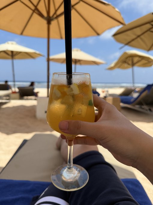 A hand raises a chilled, fruit-filled cocktail with a black straw while lounging on a beach chair, surrounded by sunlit umbrellas and the ocean in the background — perfectly embodying the carefree, breezy vibe of "lying on the beach."